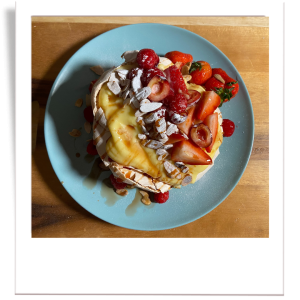 A beautifully crisp and airy Pavlova topped with creamy custard, fresh strawberries, raspberries, cherries, and toasted almond slices.