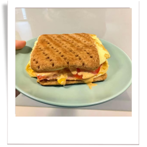 A high-protein breakfast sandwich made with whole wheat thins, fluffy folded omelette, ham, melted cheese, sweetcorn, fresh tomato slices, and a drizzle of sriracha mayo.